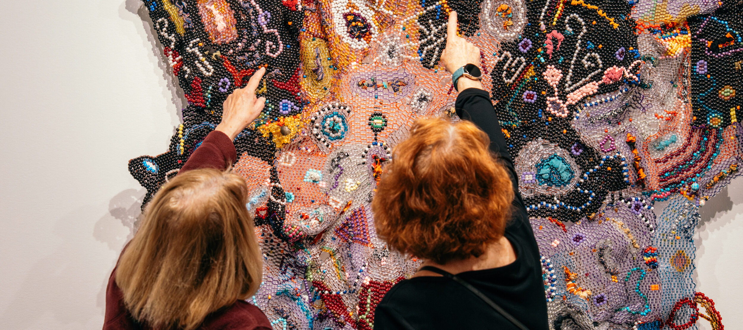 Two museum visitors gesture at a beaded work of art on the wall in a modern gallery.