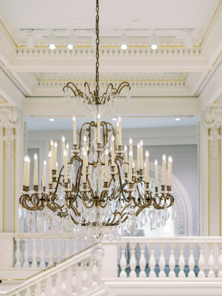 An ornate crystal chandelier with numerous candle-shaped bulbs hangs from a high ceiling in an elegant, classical interior with white railings and decorative molding.