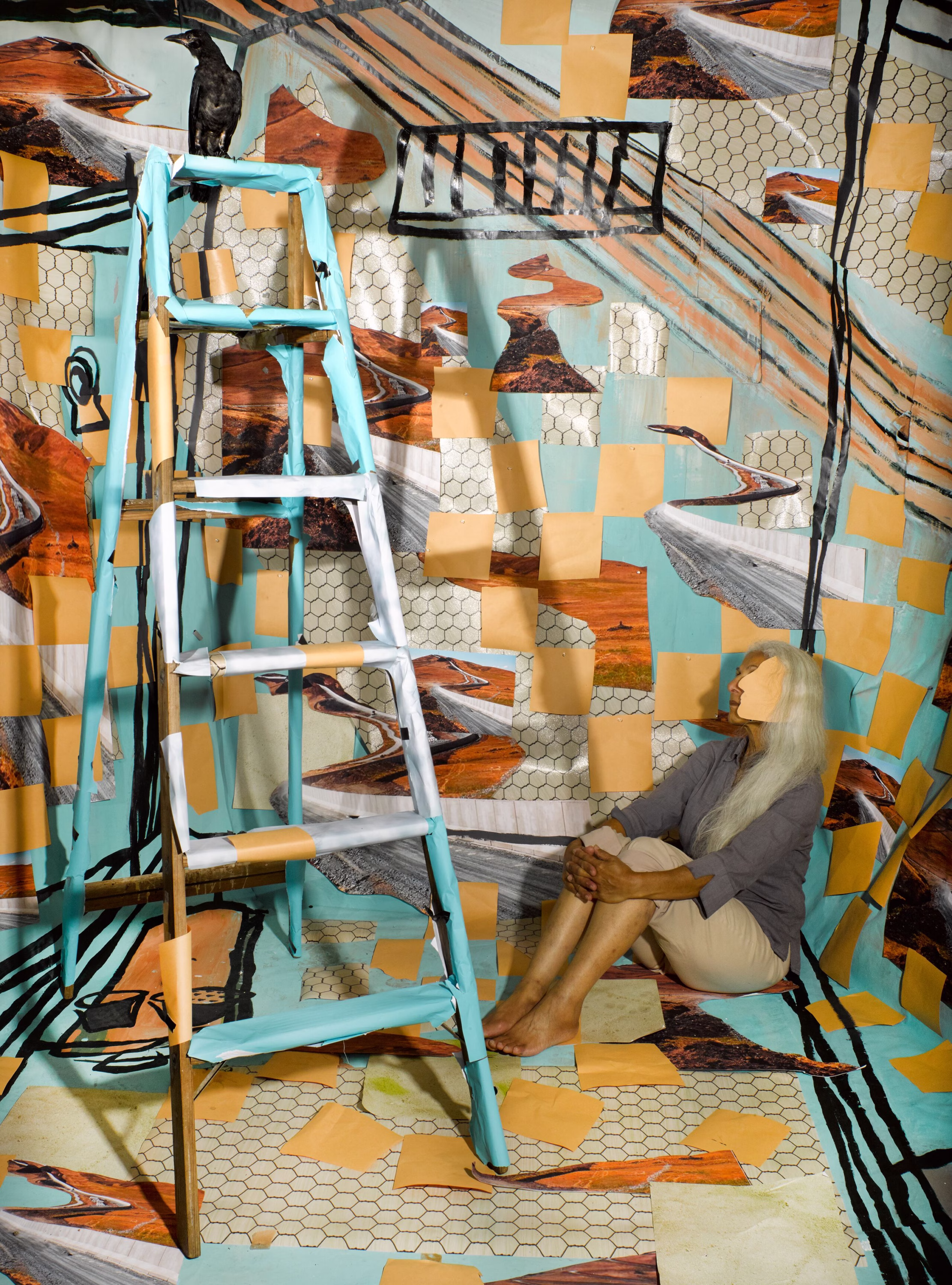 Surreal staged photograph with a masked figure seated amid layered images, patterned textiles, and cut photographic fragments forming a dense collage-like scene.