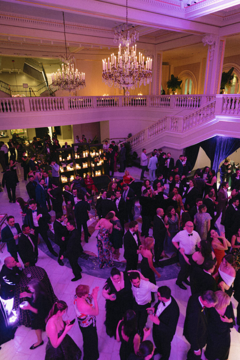 A large group of elegantly dressed people dance and mingle in a grand ballroom with chandeliers, marble floors, and a sweeping staircase in the background, under warm, colorful lighting.
