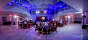 Elegant banquet hall with round dining tables and red chairs arranged on a shiny white floor, dramatic purple lighting, chandeliers overhead, and grand staircases on either side, set for a formal event.