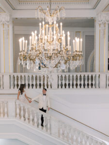 A couple dressed in elegant wedding attire holds hands while descending a grand white staircase in a lavish hall, illuminated by a large ornate chandelier above them.