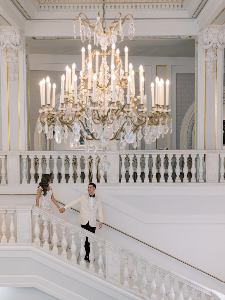 A couple dressed in elegant wedding attire holds hands while descending a grand white staircase in a lavish hall, illuminated by a large ornate chandelier above them.