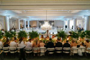 A large group of people dressed formally are seated at a long, elegant dining table with lush green centerpieces and candles in a grand, ornate hall with high ceilings and a large chandelier.