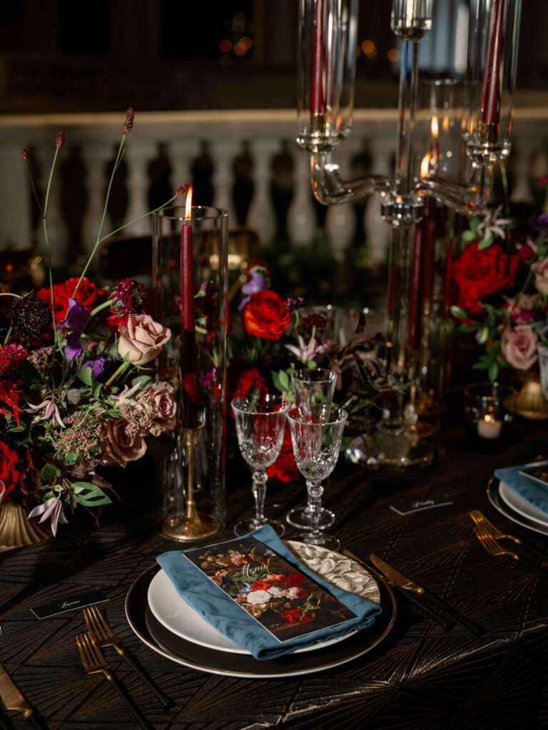 Elegant dinner table set with dark floral arrangements, tall red candles in glass holders, crystal glasses, gold cutlery, and a napkin with a floral design on a black plate and blue napkin, on a dark textured tablecloth.