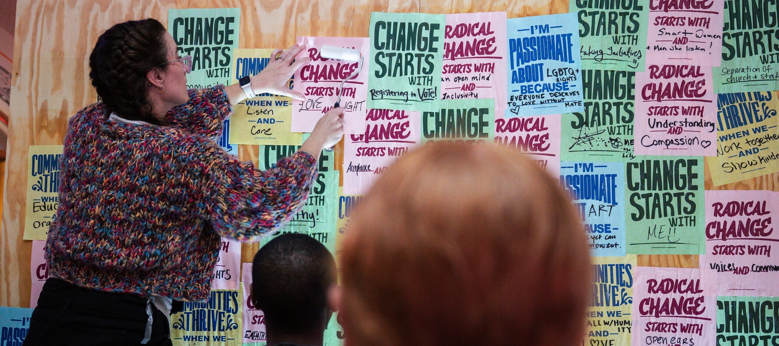 A woman pastes a paper with the text “Radical change starts with me!” onto a wall covered in colorful, handwritten posters with motivational messages, while two people look on from the foreground.