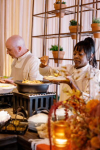 Two people serve themselves food from buffet dishes on a table, surrounded by decorative plants and autumn-themed decor. Both are dressed in light-colored clothing and focused on making their plates.
