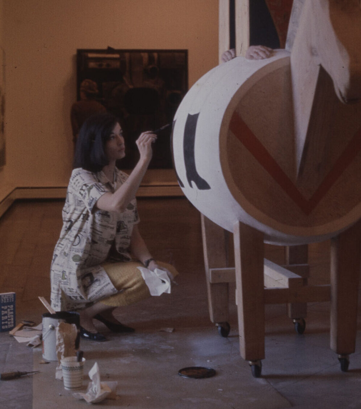 A color photograph of a woman with light skin and brown hair crouching indoors next to a large wooden sculptural artwork, painting it with black paint.