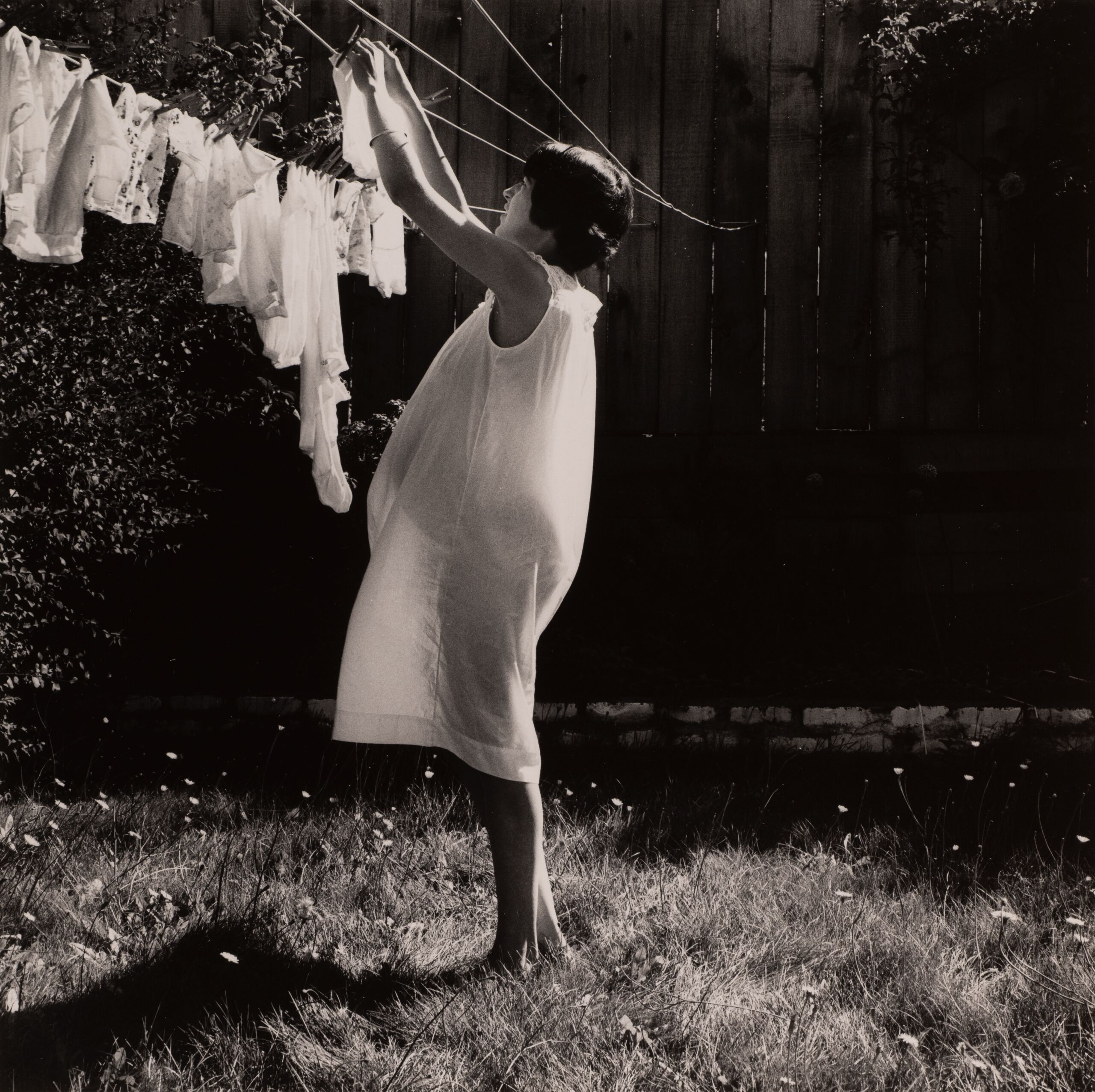 A black and white photo of a pregnant woman in a white dress hanging clothes on a line in a backyard.