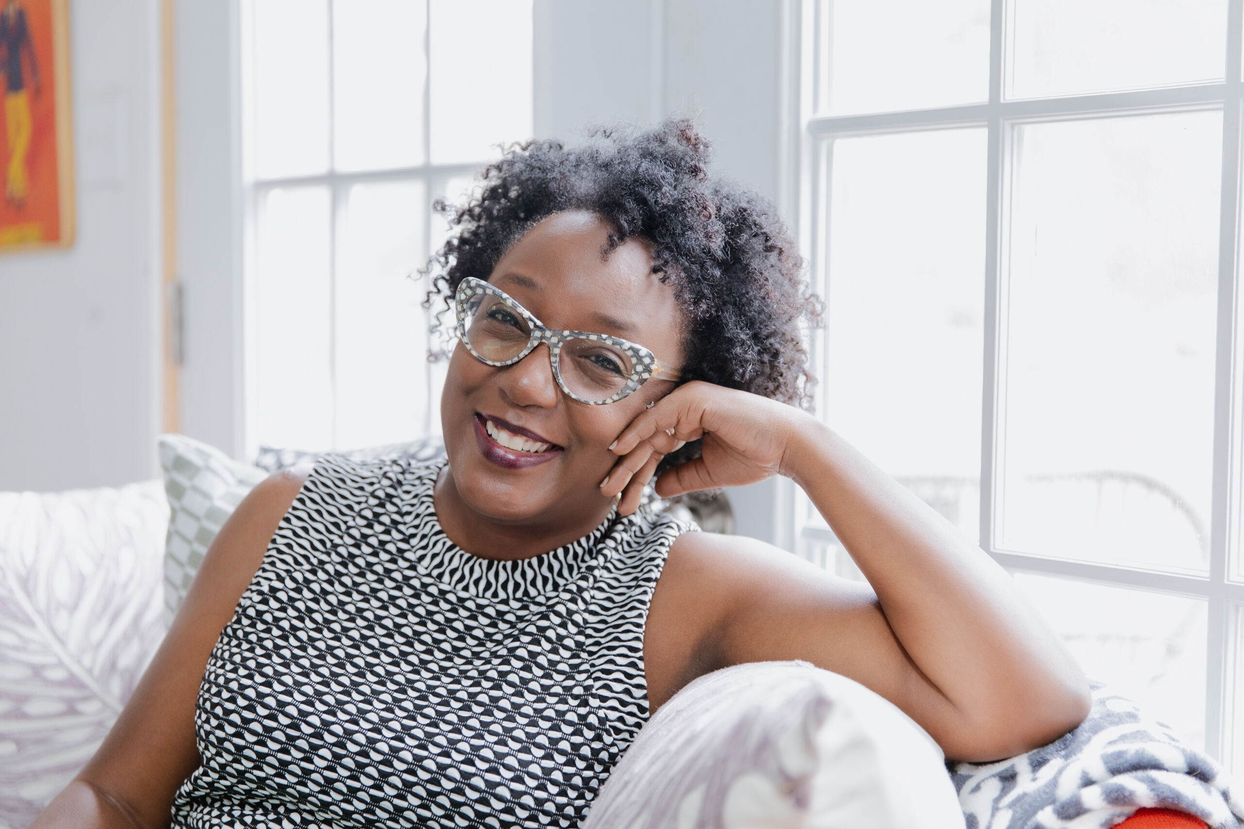 A woman with dark skin tone and short, black, curly hair smiles and wears bold gray-and-white polka dot glasses and a sleeveless top with a black-and-white geometric pattern.