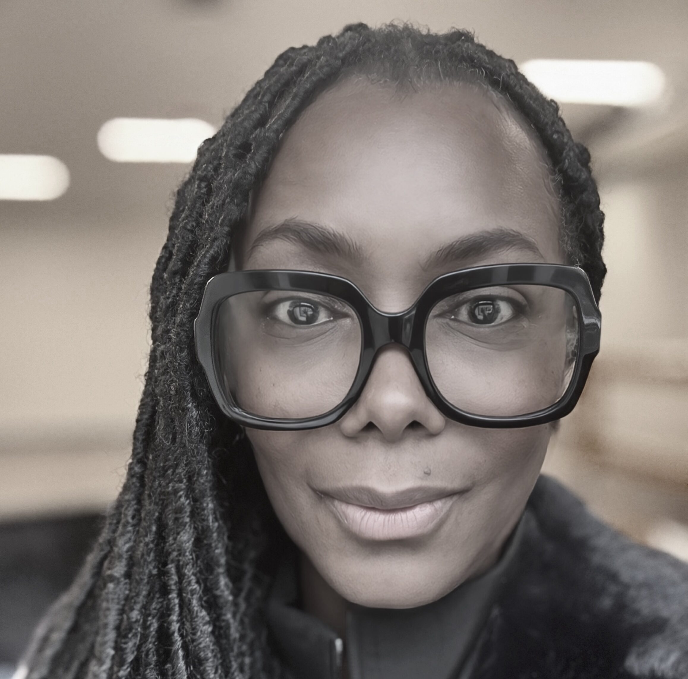 A photograph of a woman in black and white. She has a medium skin tone and is wearing big, black glasses.
