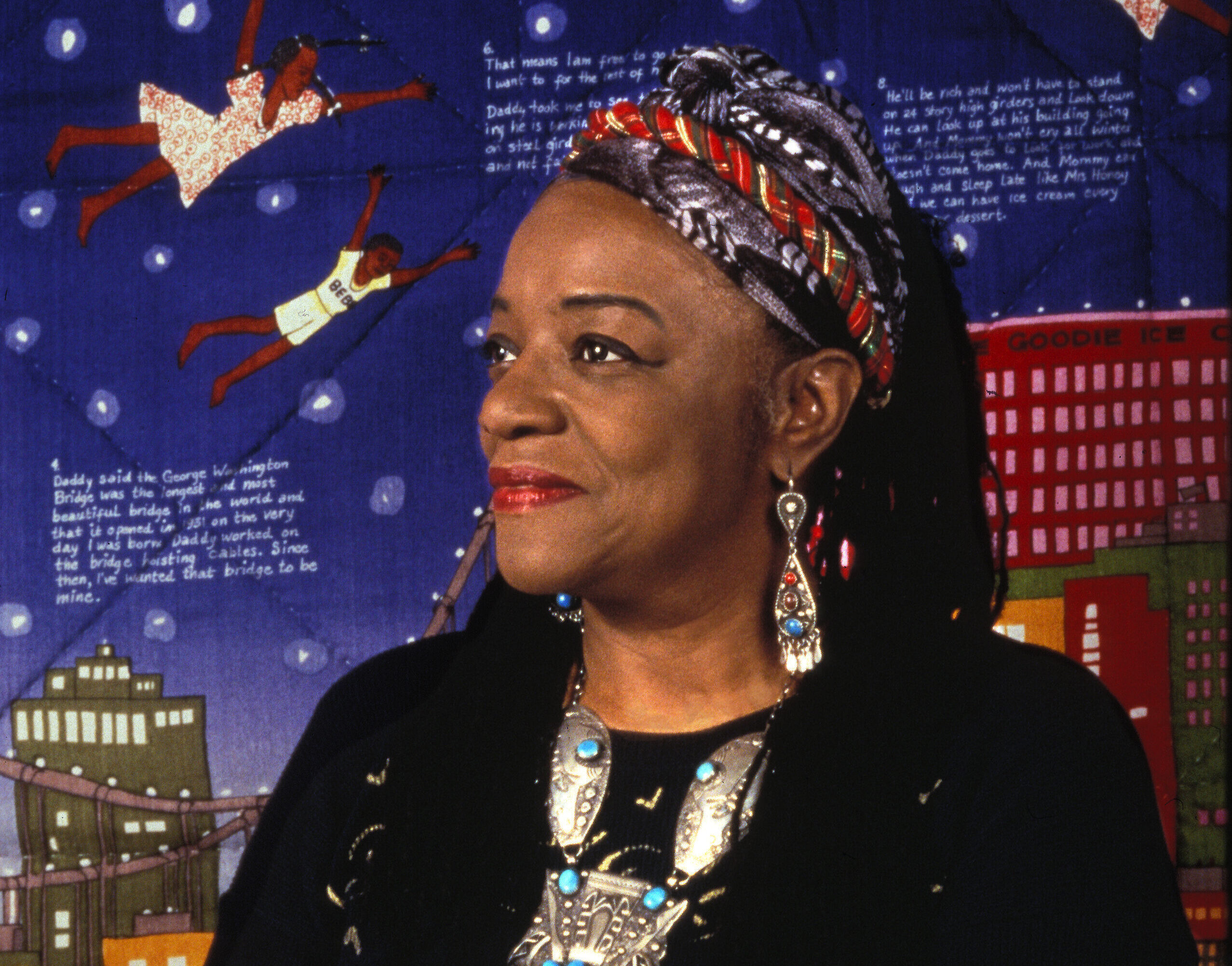 A middle-aged woman with dark skin tone wears a black long-sleeved top and colorful headscarf over her black hair.