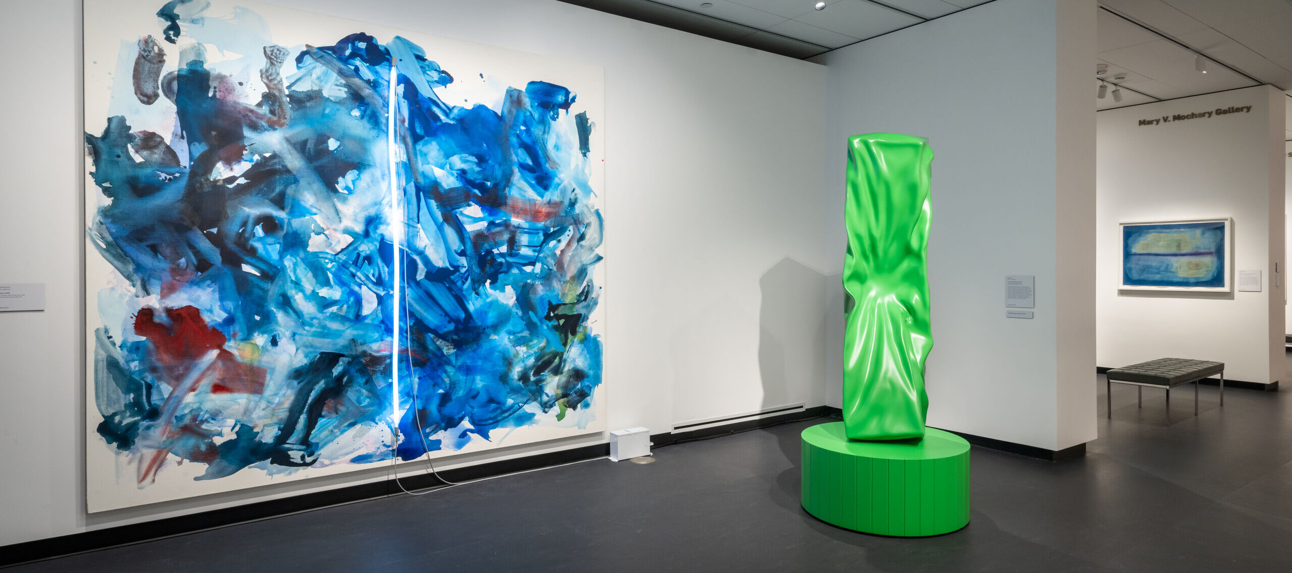 View of an indoor art museum gallery space. A large abstract artwork consisting of blue, red, and green paint and a fluorescent rod in the center hangs on the wall on the left. A green sculpture of a rectangular column that looks like it was punched sits on a platform to the right. In the background hangs a blue abstract painting.