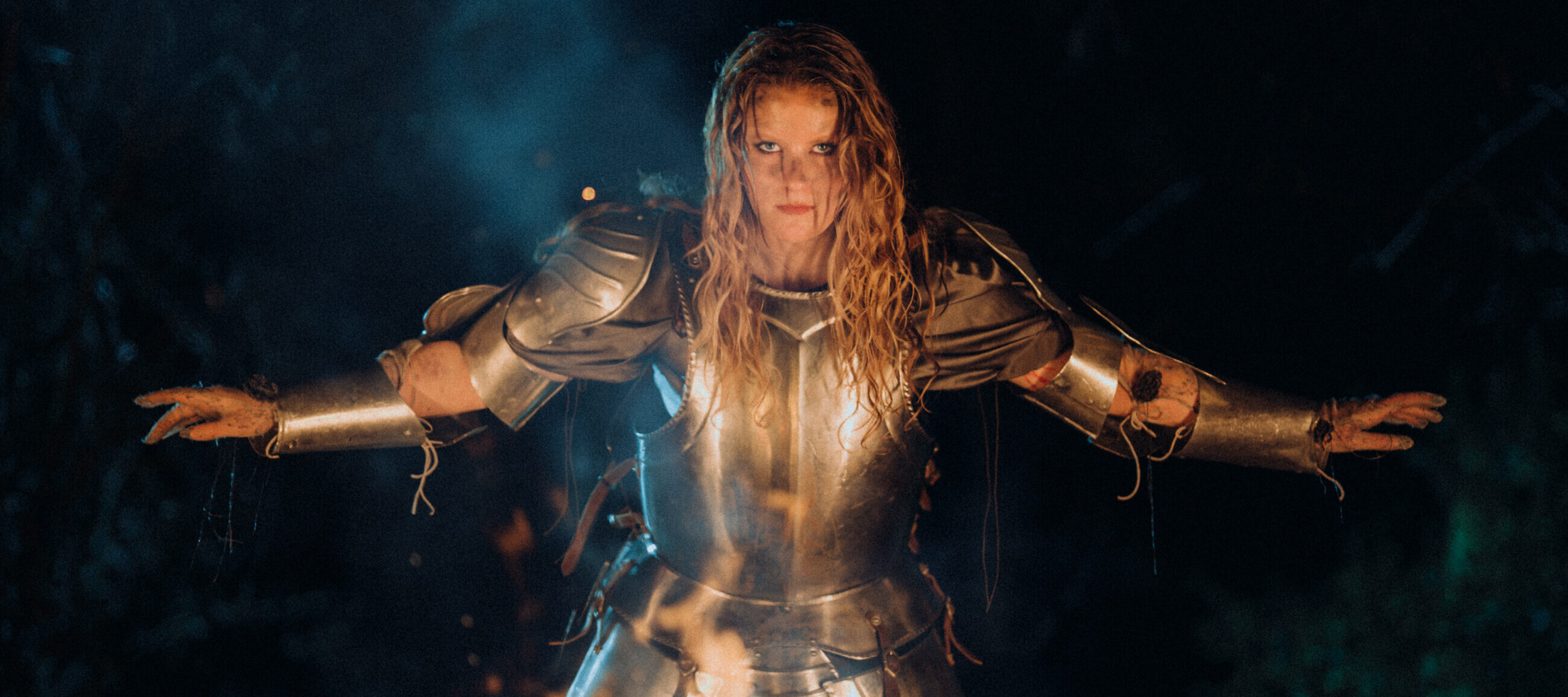 A person with long, wet hair wearing shiny metal armor stands with arms outstretched in front of a fire at night, illuminated by flames and surrounded by smoke and darkness.