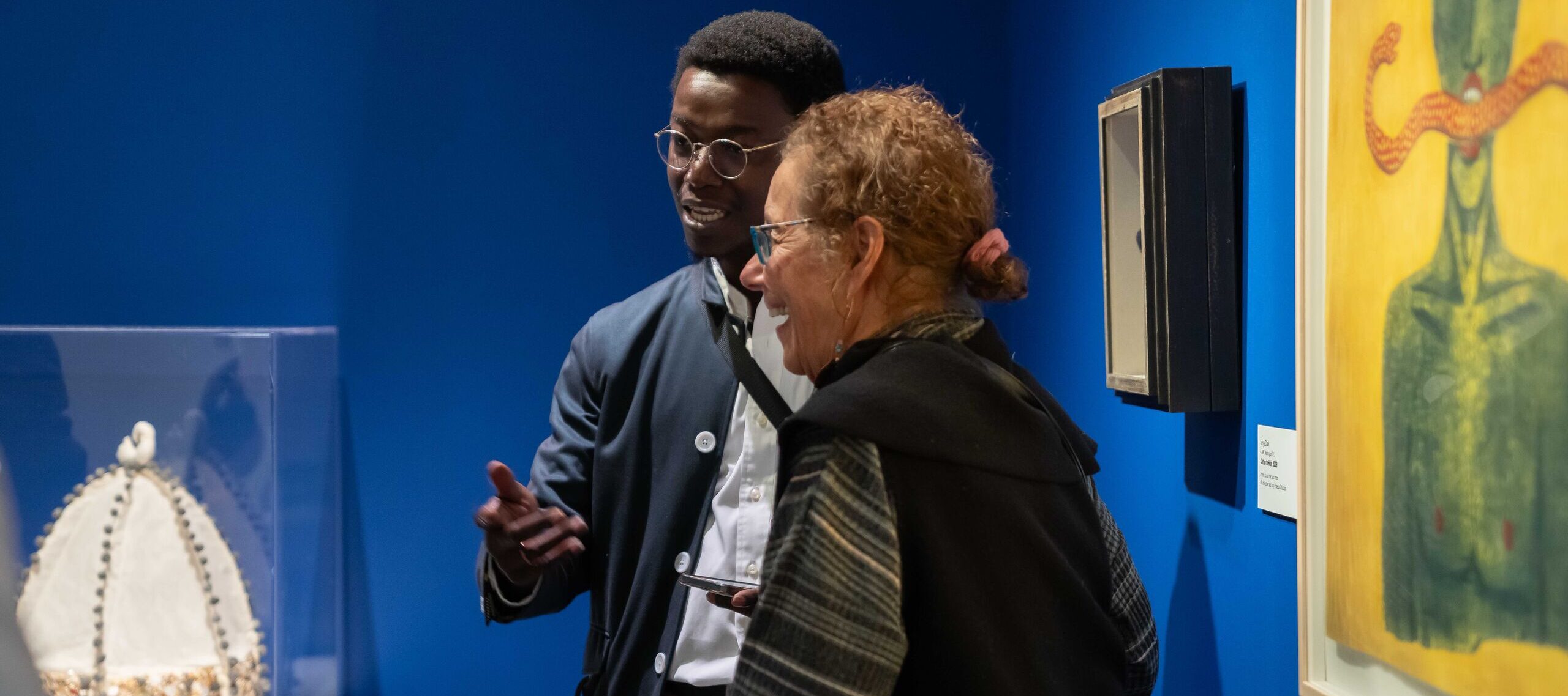 Two museum visitors in conversation next to an art piece. A man with a dark skin tone, glasses with a golden frame wearing a navy blazer is talking to a woman with a medium-light skin tone wearing a black jacket.