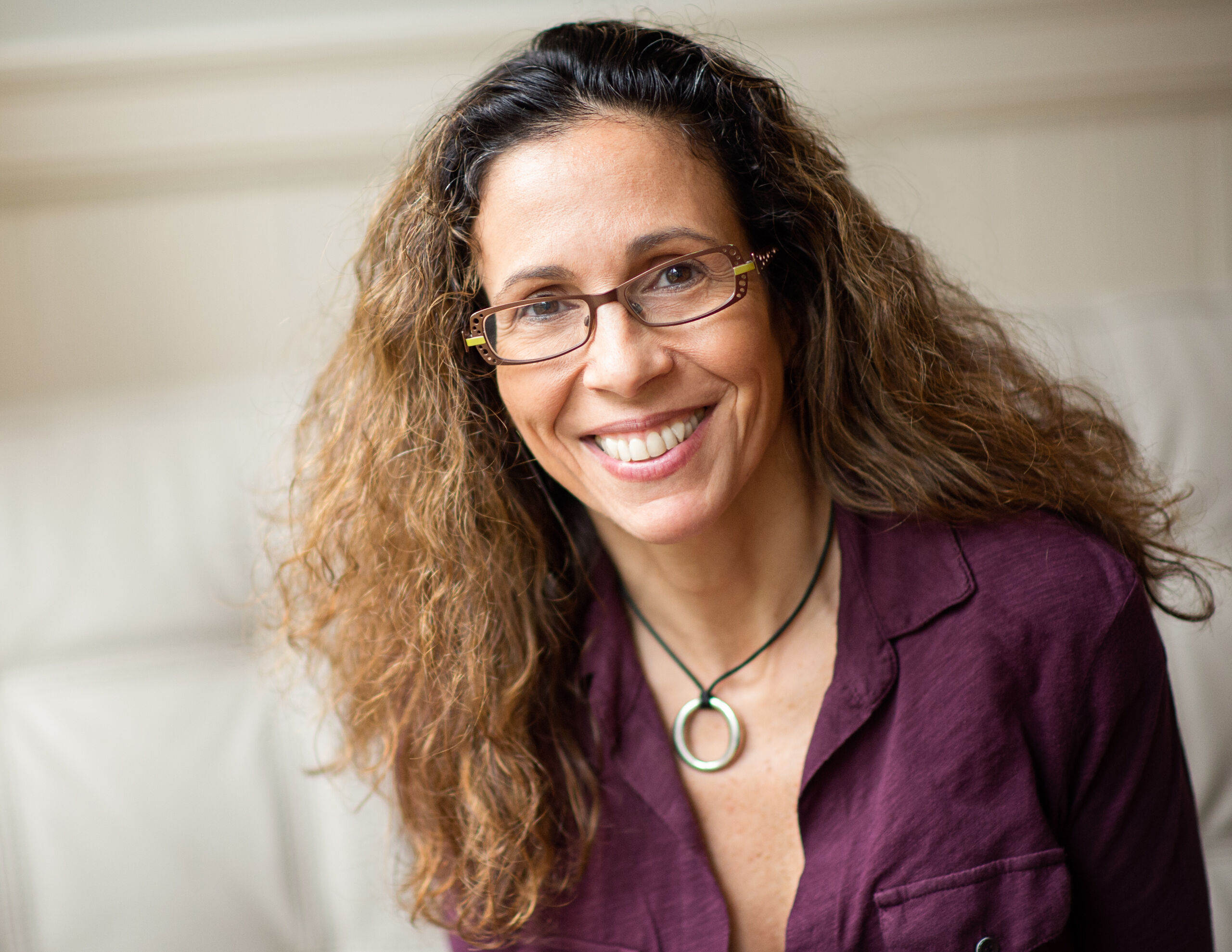 A photographic portrait of a woman with a medium-light skin tone and curly, brown hair. The woman wears a purple blouse, glasses, and a chunky silver necklace. She sits before a white background and smiles at the camera.