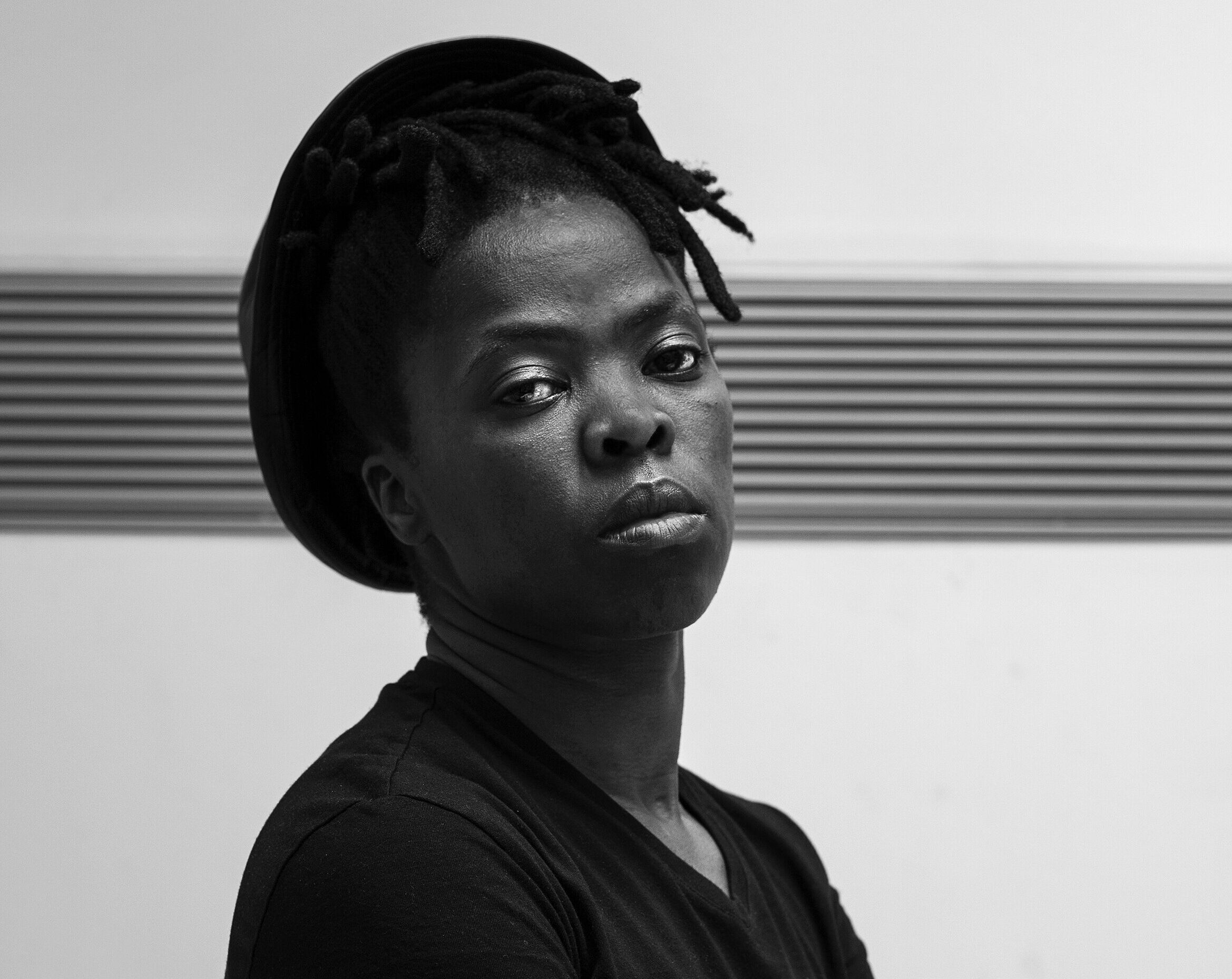 A black and white photo of a dark-skinned person crossing their arms and wearing a black t-shirt and brimmed hat.