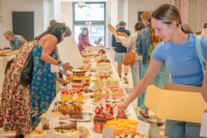 People select cakes and desserts from long tables covered with a variety of colorful baked goods, while holding boxes in a large bright hall with a glass door at the end.