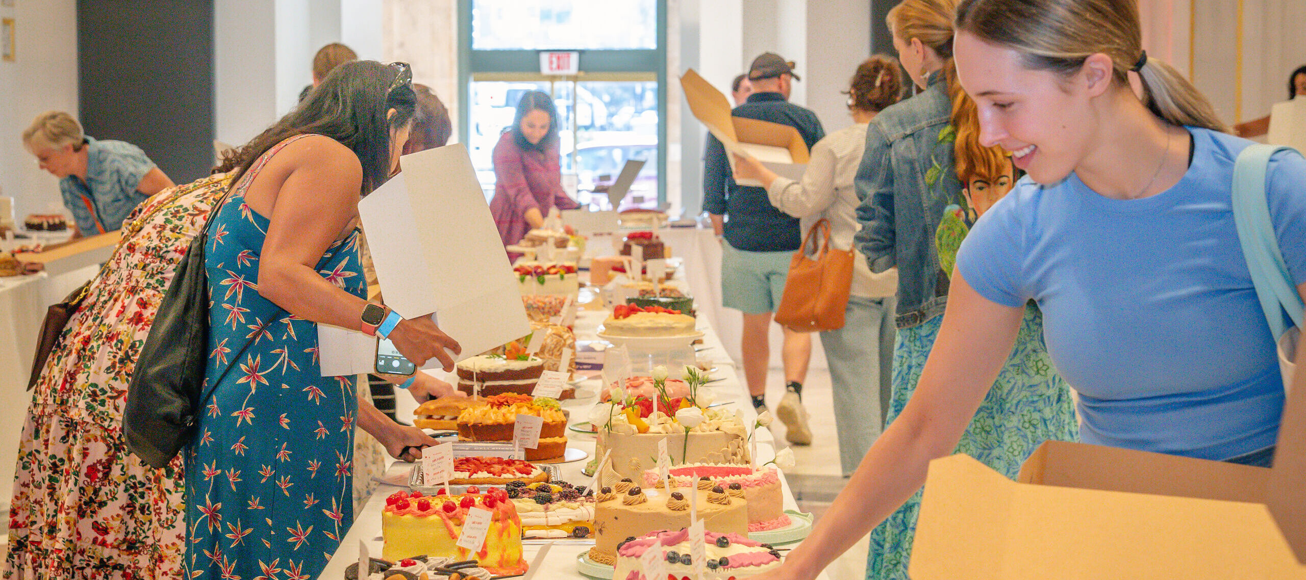 People select cakes and desserts from long tables covered with a variety of colorful baked goods, while holding boxes in a large bright hall with a glass door at the end.