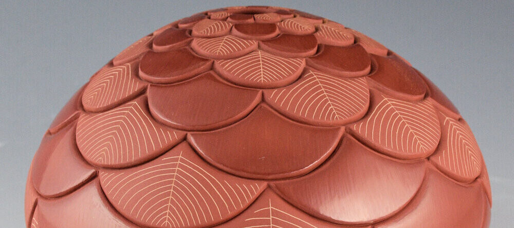 A reddish-brown ceramic vessel with a dome shape, decorated with carved overlapping petal-like scales and fine etched line patterns, creating a textured, layered appearance.