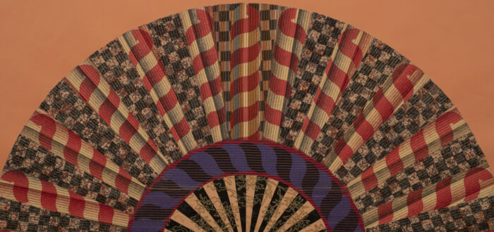A colorfully painted and collaged folding fan spread open on a peach-orange background.