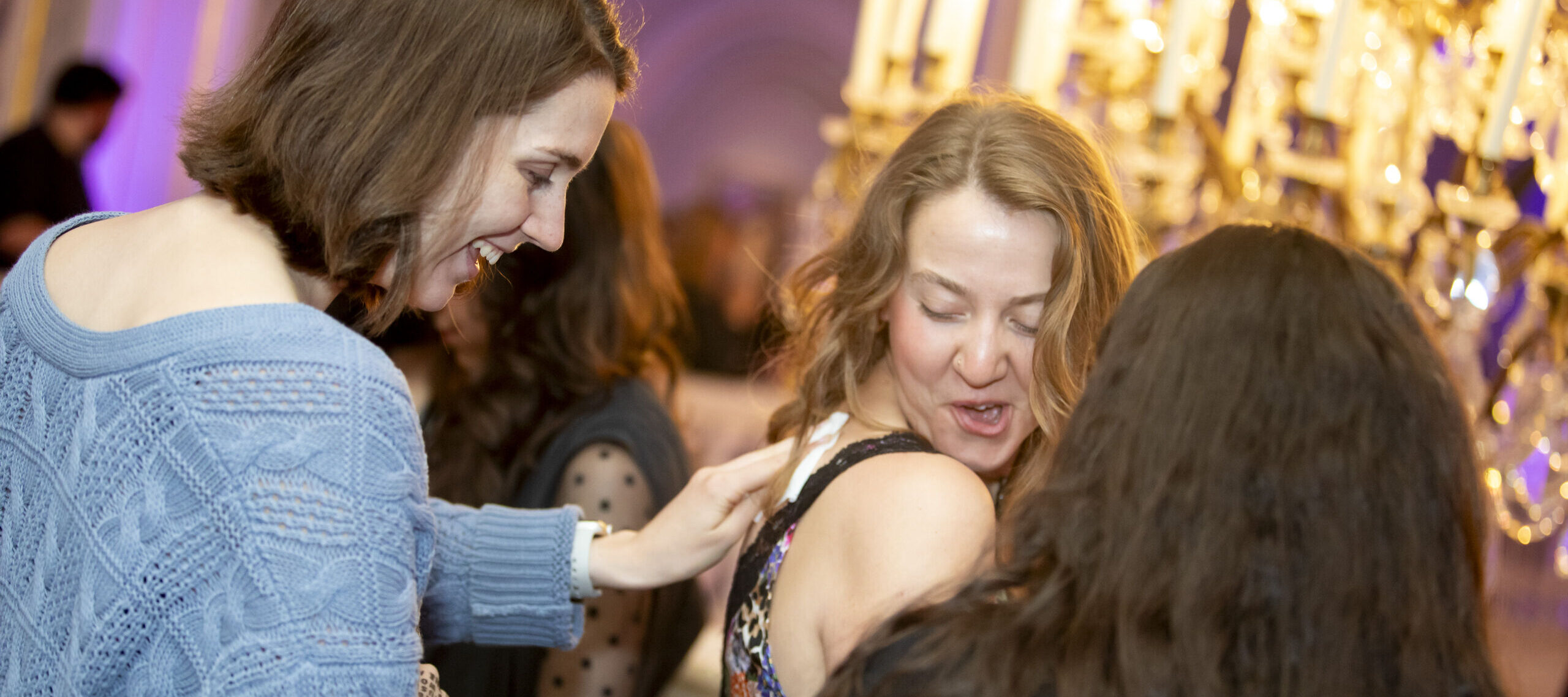 Three women are laughing and interacting at a lively indoor event, with warm lighting from a chandelier in the background. One woman in a blue sweater is touching another woman’s shoulder.
