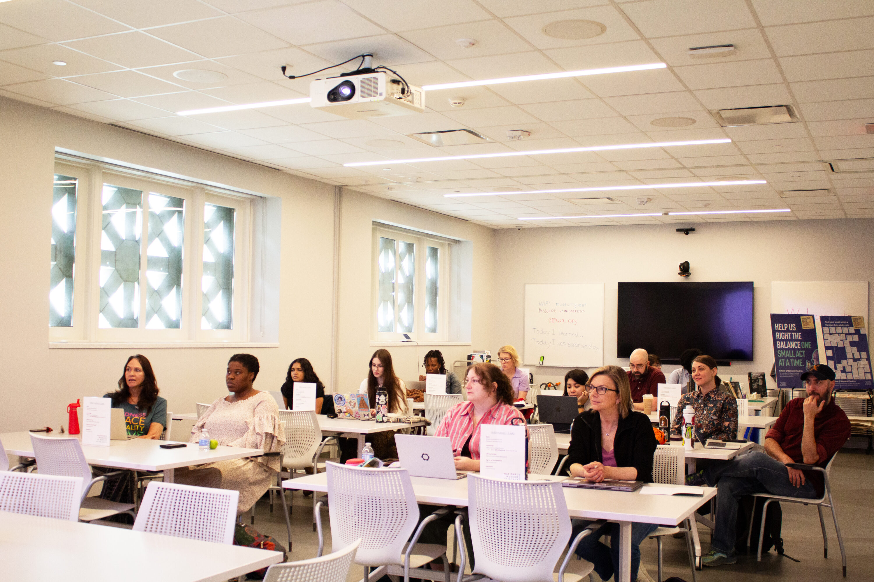A diverse group of adults seated at white tables in a modern classroom, attentively facing the front. A projector hangs from the ceiling, and a whiteboard and TV screen are visible at the back of the room.