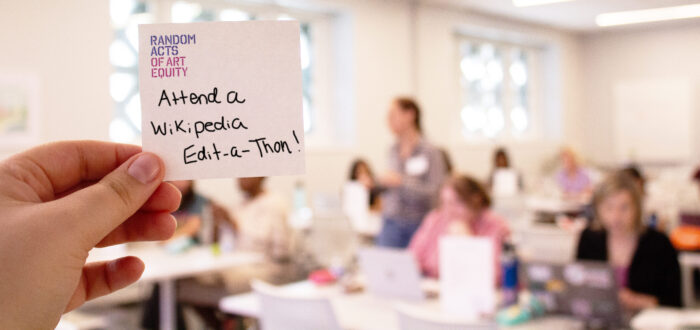 A hand holds a sticky note reading “Attend a Wikipedia Edit-a-Thon!” in a bright classroom where people are gathered, working on laptops in the background.