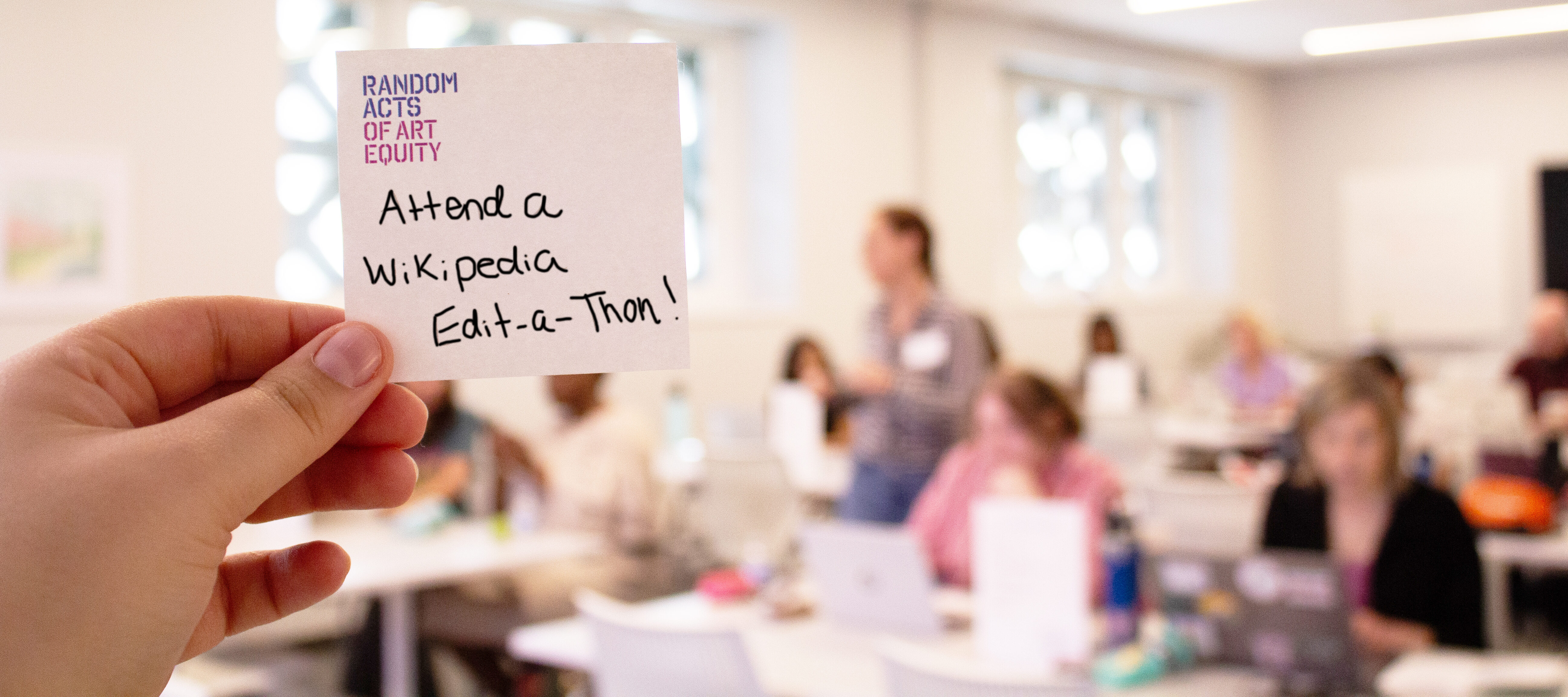 A hand holds a sticky note reading “Attend a Wikipedia Edit-a-Thon!” in a bright classroom where people are gathered, working on laptops in the background.