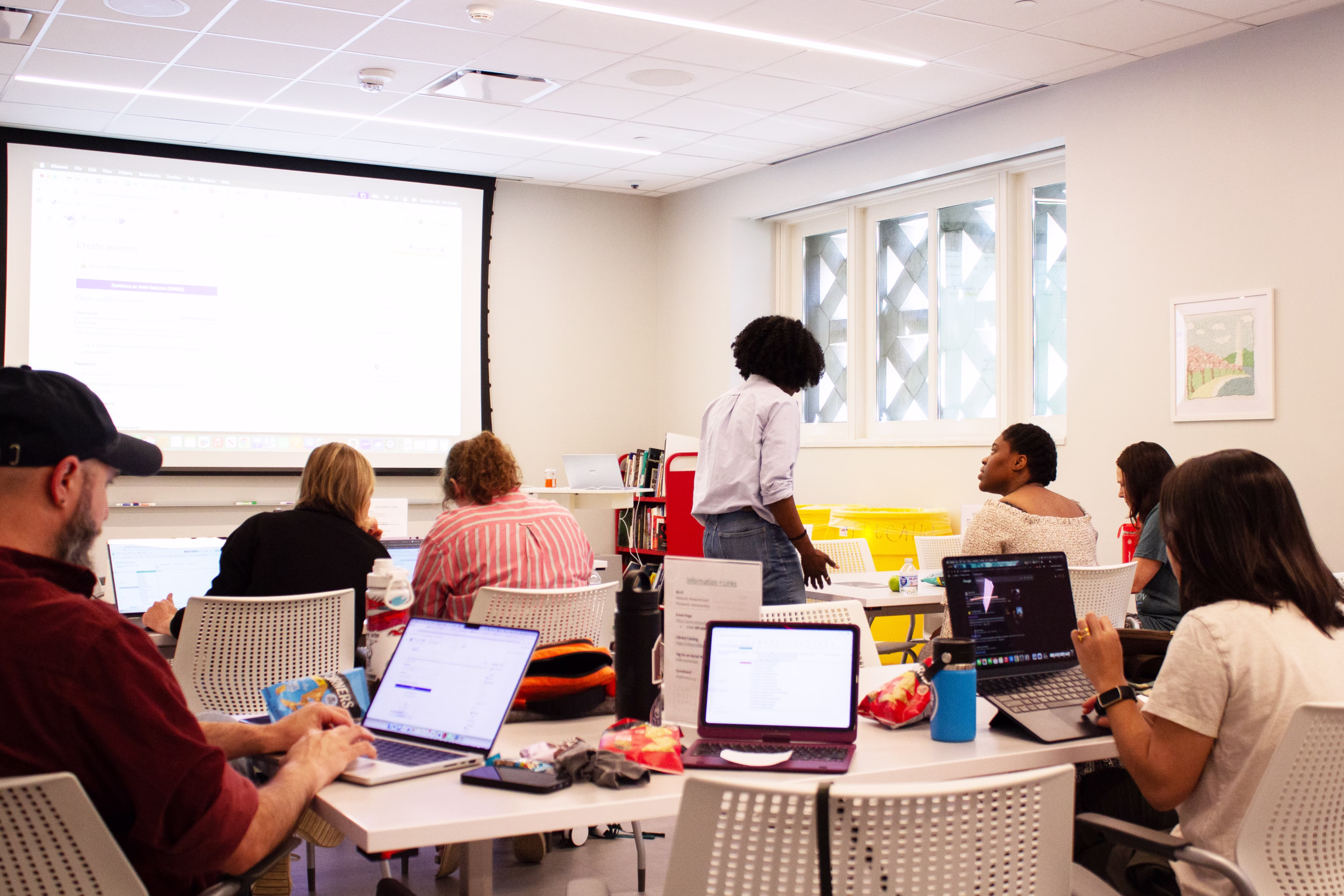 People sit at tables in a bright classroom, using laptops and watching a presentation projected on a screen at the front of the room. One person stands, while others are seated and engaged with their devices.