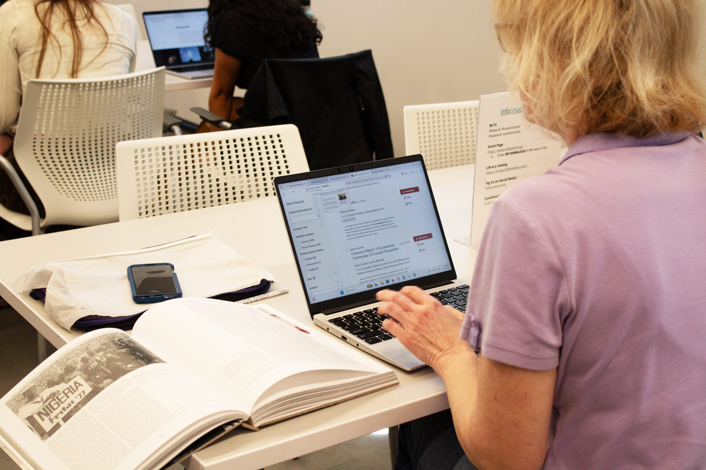 Seen from the back, a woman sits at a desk with a large book open and works on her laptop.