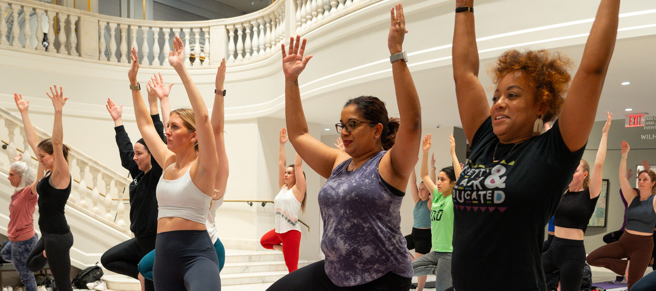 A crowd participates in a yoga class in the Great Hall of the National Museum of Women in the Arts. They have their arms raised above their heads and one foot propped on their opposite leg in a "tree" pose.