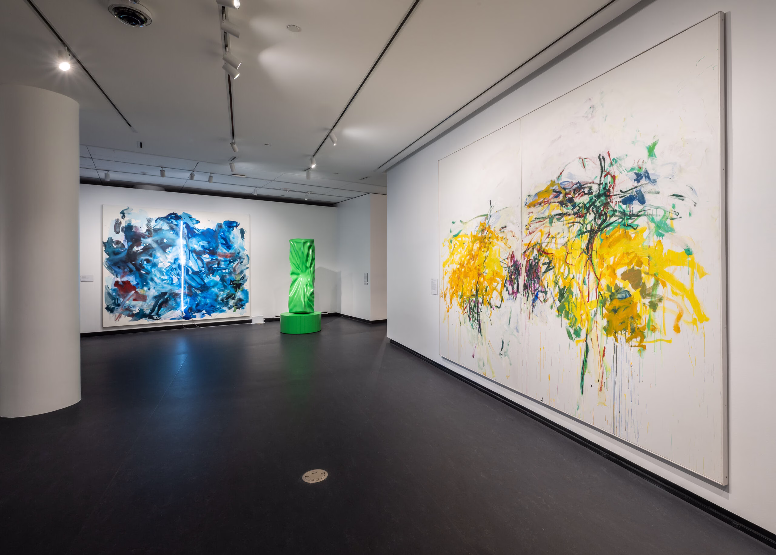 A modern museum gallery with large abstract paintings on white walls; one features yellow and purple splashes, another blue and white swirls, and a green sculptural piece stands in the center of the dark-floored room.