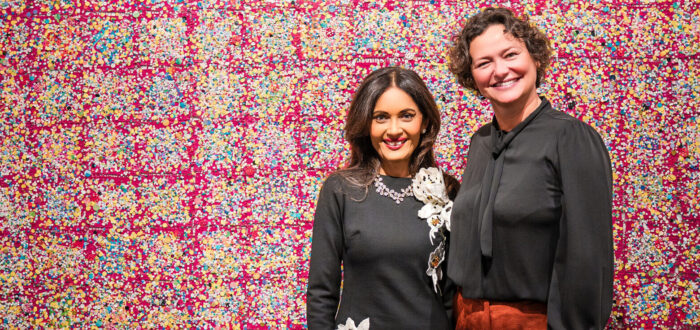 Two women stand smiling in front of a large, colorful abstract artwork with a red, pink, and yellow grid pattern. One woman wears a black dress with floral designs; the other wears a black blouse and rust-colored pants.
