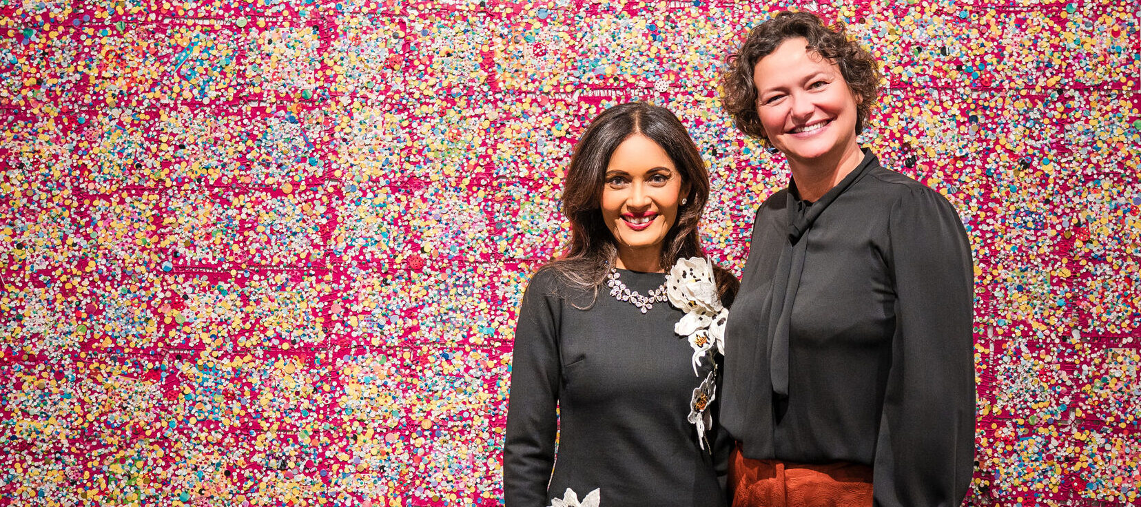 Two women stand smiling in front of a large, colorful abstract artwork with a red, pink, and yellow grid pattern. One woman wears a black dress with floral designs; the other wears a black blouse and rust-colored pants.