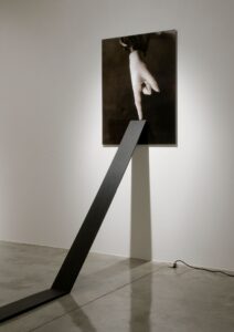 A black and white photograph of a hand with pointer finger extended displayed on a wall with a wooden board leaning against it at an angle.