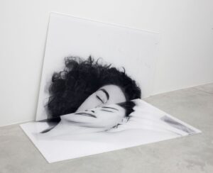A black and white photograph of a woman's head, divided in two, with one half resting against the wall and one half on the floor.