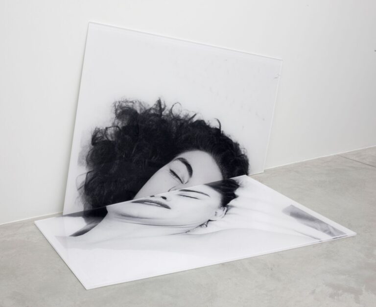A black and white photograph of a woman's head, divided in two, with one half resting against the wall and one half on the floor.