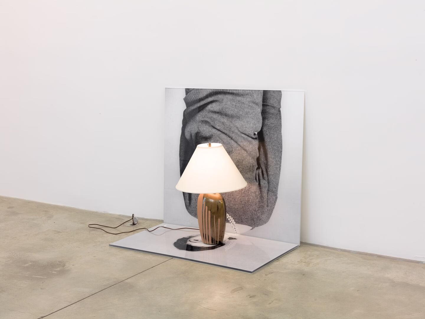 A black and white photograph of a woman's head and torso, divided in two, with one half resting against the wall and one half on the floor. A table lamp rests on top of her face on the floor.