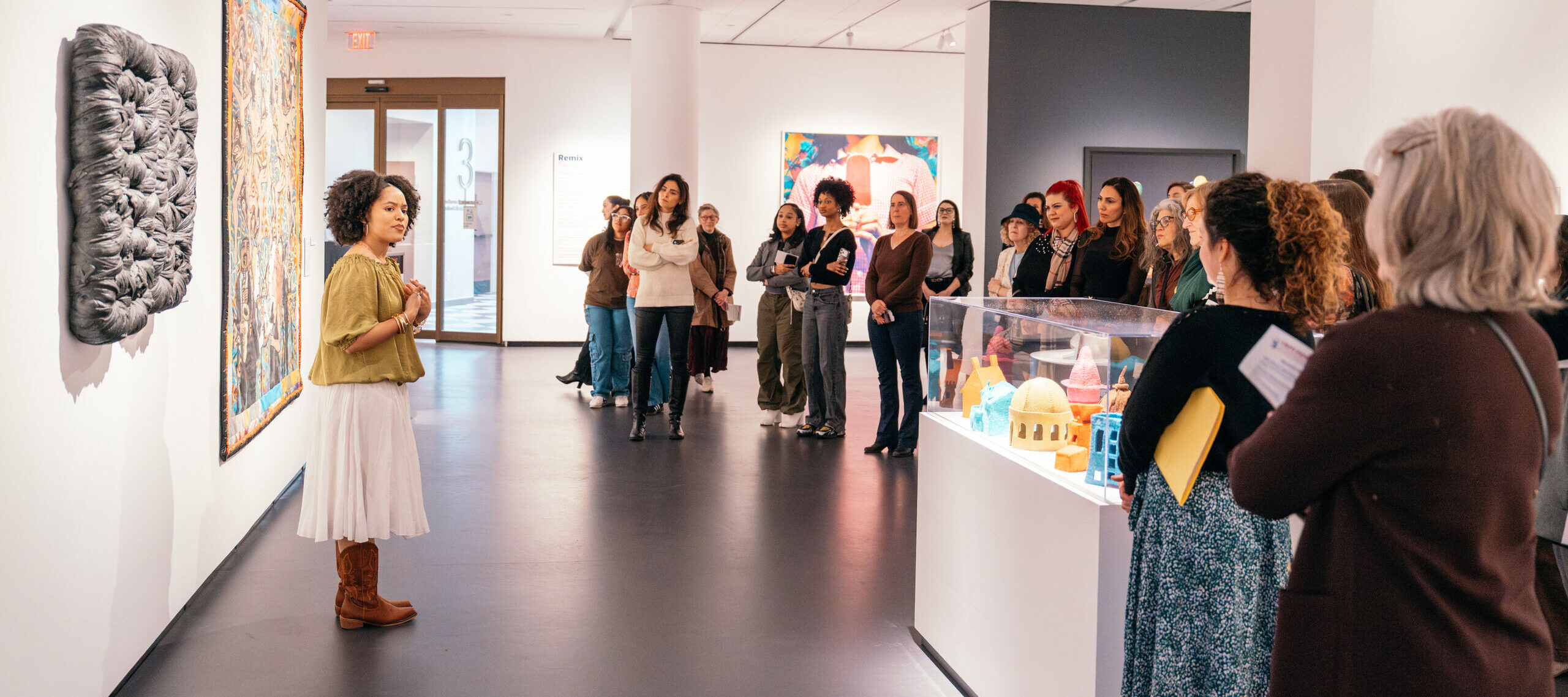 A woman speaks to a group of people in an art gallery. The group listens attentively, surrounded by modern artwork on the walls and display cases. The setting is bright with white walls and spacious walkways.