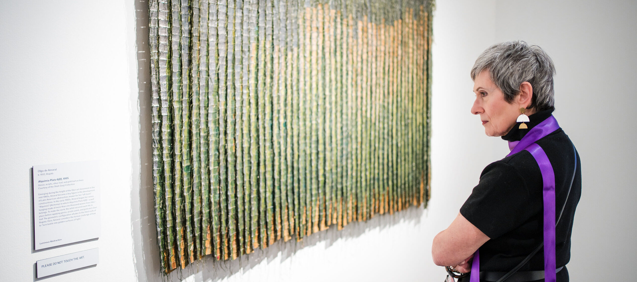 A woman stands with arms crossed, closely observing a textured, vertically striped woven artwork displayed on a white gallery wall. A descriptive placard is visible beside the piece.