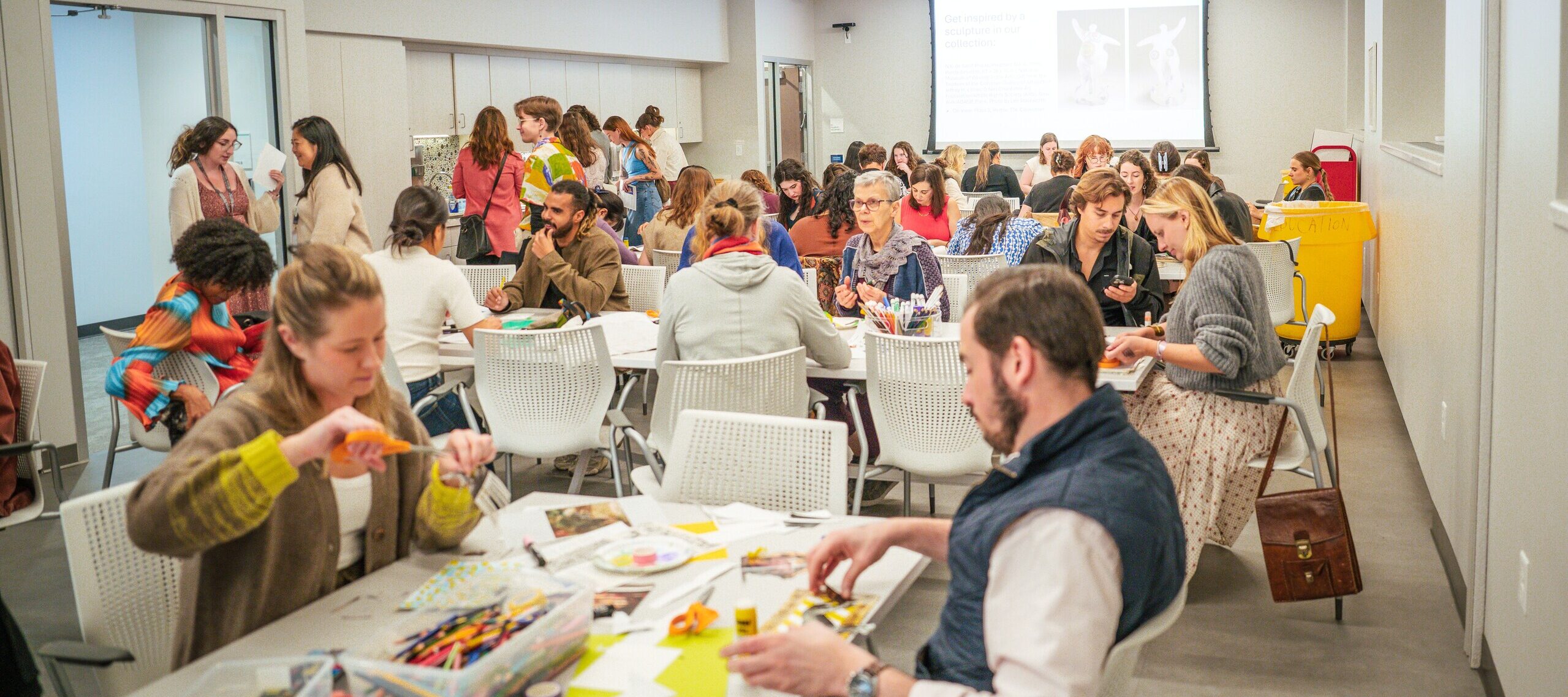 A large group of people sit at tables in a bright, modern classroom, working on arts and crafts projects with colorful supplies. Others stand and talk in the background. A presentation is displayed on a screen.