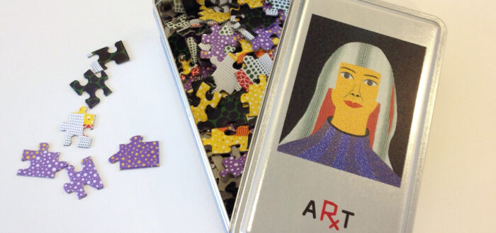 A metal tin with a colorful jigsaw puzzle inside is open on a white surface. The tin's lid displays an illustration of a woman with long hair and a blue top, labeled "ART." Some puzzle pieces are scattered outside the tin.