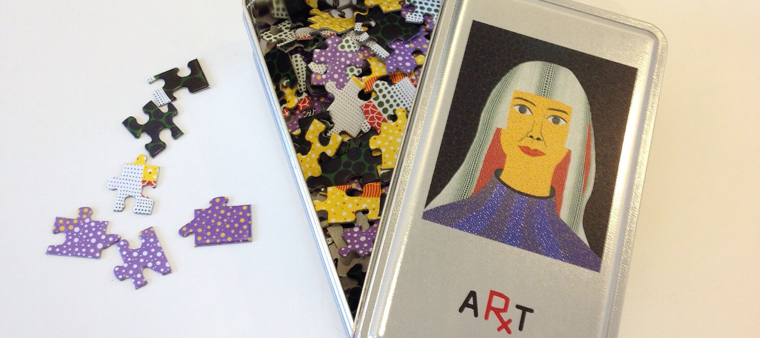 A metal tin with a colorful jigsaw puzzle inside is open on a white surface. The tin's lid displays an illustration of a woman with long hair and a blue top, labeled "ART." Some puzzle pieces are scattered outside the tin.