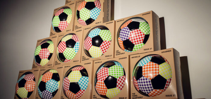 A pyramid stack of boxed soccer balls with colorful checkered patterns on each panel, displayed against a plain wall with light and shadow effects.