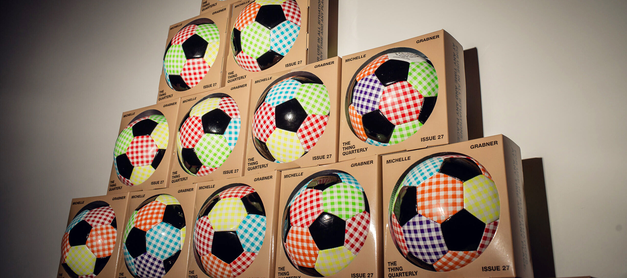 A pyramid stack of boxed soccer balls with colorful checkered patterns on each panel, displayed against a plain wall with light and shadow effects.
