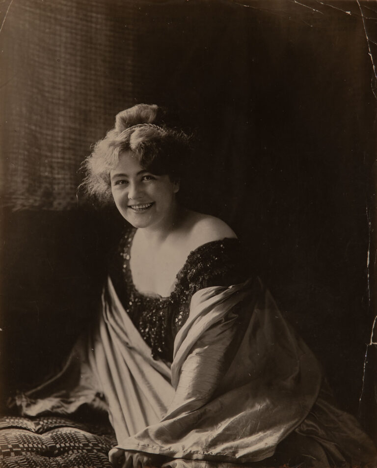 A woman with light skin tone smiles out at the viewer from a dramatically lit black-and-white photograph. Her wavy fair hair is pulled back in an old-fashioned style, and she wears a dark gown with sequins and ruffles as well as a silk wrap.