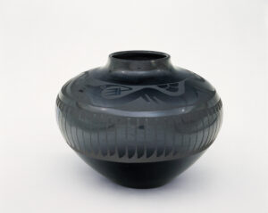 A blackware pottery jar has a small mouth and widens into a rounded shape in the middle, tapering at the base. The shape of a serpent with jagged sharp teeth and pronged horns is outlined in a matte coating set against the glossy finish. Below the serpent is a line of repeating, upright feather shapes that point toward the bottom of the jar.
