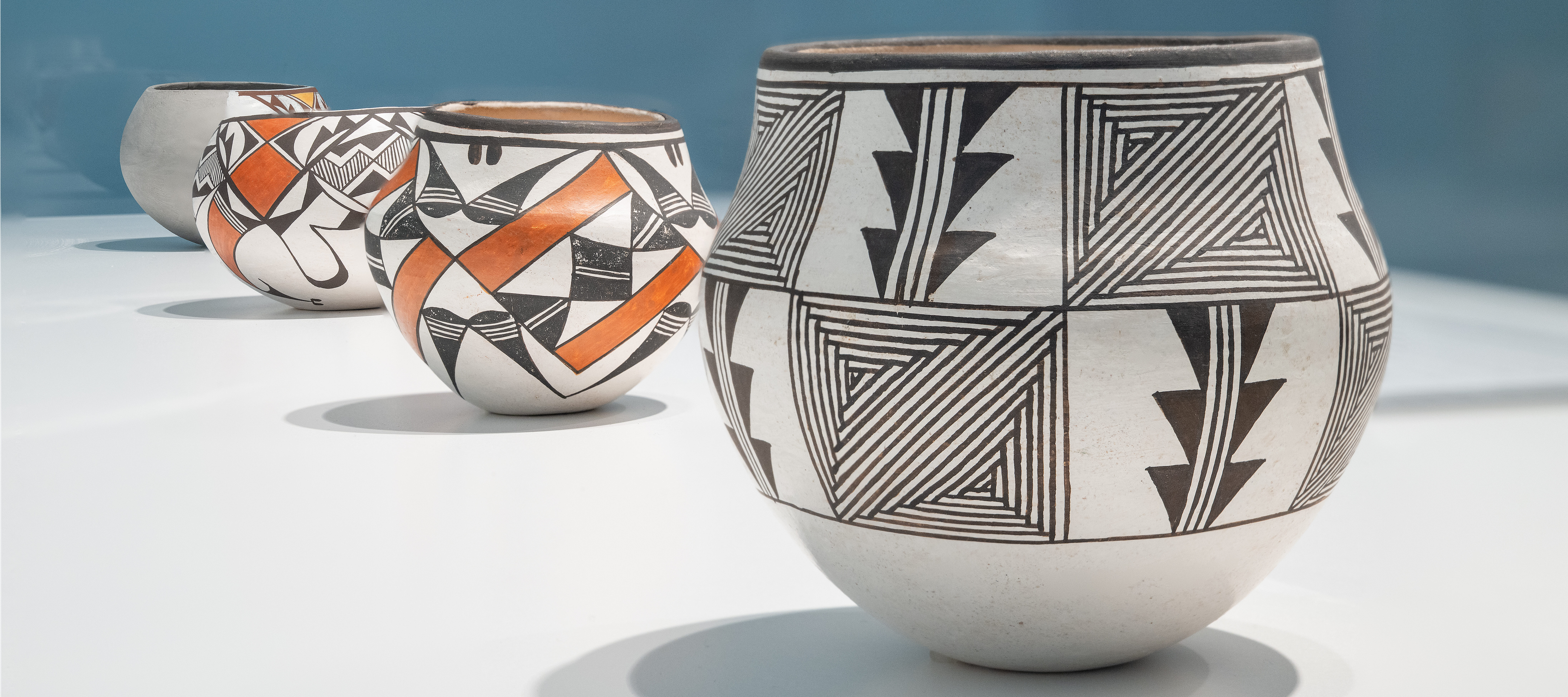Four ceramic pots with intricate black, white, and red geometric patterns sit in a row on a white surface against a blue background. The pots feature various designs.