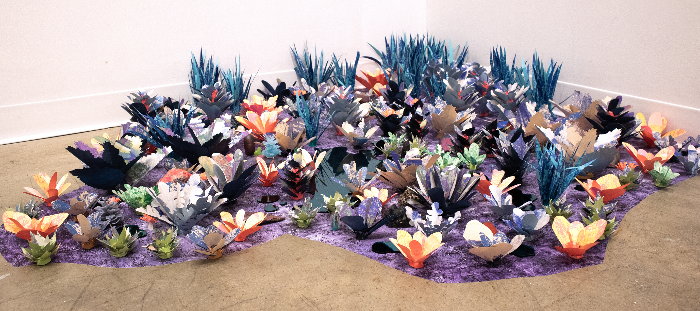 A sprawling field of multicolored paper and vinyl plants and flowers on a purple surface in the corner of a gallery.